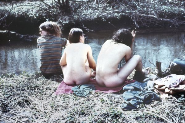 Rear view of three people, two nude and one clothed. They are sitting on a towel next to a pile of clothes on the bank of Rowan Creek near York Farm during the Sound Storm music festival.