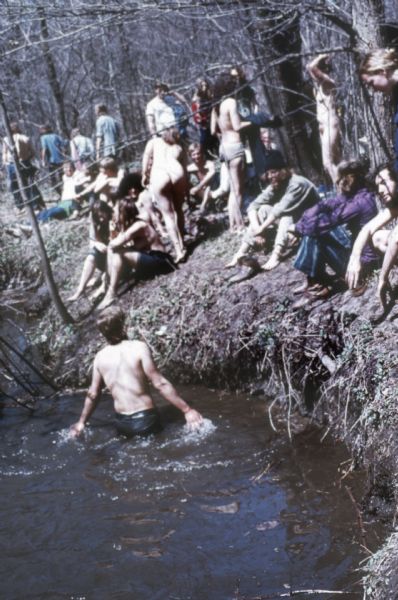 A group of young men and women, some nude, sitting on the bank of Rowan Creek near York Farm during the Sound Storm music festival.