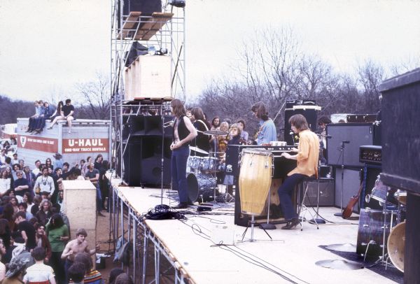 Elevated view of The Bowery Boys performing onstage at the Sound Storm music festival. Band members are Ken Heim on organ, Richard Wiegel on guitar, Jeff Amundson singing, Cubby Tracy on drums, and Steve Tracy on bass. A group of people are sitting on a U-Haul truck on the left, and stage scaffolding is on the left corner of the stage. A crowd is in front of the stage.