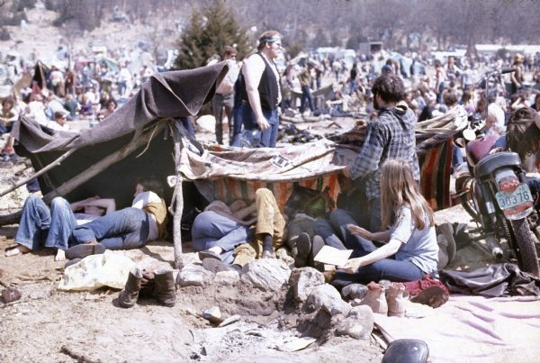 Members of the Sound Storm music festival audience crowd under improvised tents partially supported by the handlebars of a motorcycle and tree limbs. There is a fire pit in the foreground. The camp extends up the hill in the background.
