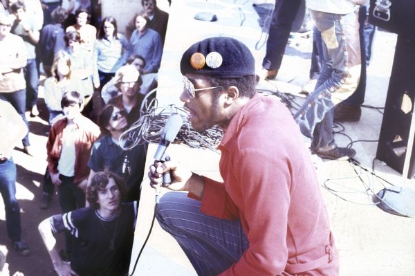 Unidentified male African American artist, wearing a beret with two illegible buttons on it, kneels on the edge of the stage talking or singing into a hand-held microphone. Part of the audience crowds the stage on the lower left.