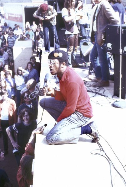 Unidentified African American male artist, wearing a beret with two illegible buttons on it, kneels on the edge of the stage talking or singing into a hand-held microphone. One audience member is holding a recording device on the front of the crowded stage.