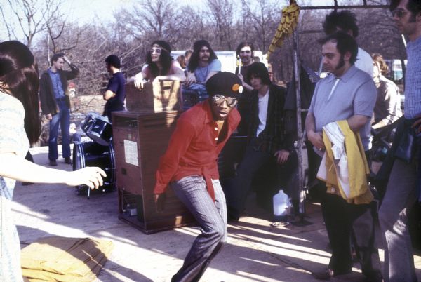 A man wearing a beret with two buttons on it dances across the stage. An audience member on the left is also dancing, and stagehands and photographers crowd the rear of the stage area. In the background, the band The Tayles lean on equipment. Left to right: Scott Eakin (wearing headband), Jeremy Wilson, Ken Ross, and Len Epand.