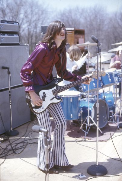 Bassist Bob Collins (with a peace symbol on his instrument) from the band Tongue performs at the Sound Storm music festival with drummer Dick Weber visible in the background. Keyboardist Mickey Larsen is partially visible further back.