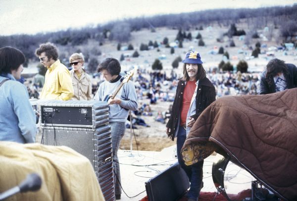 Peter Obranovich (better known at the time as "Pete Bobo"), promoter for the festival, wearing a blue and yellow bucket hat, standing on the side of the stage with other festival staff while a band prepares to play the Sound Storm music festival. A crowd is spread out on the hill behind him.