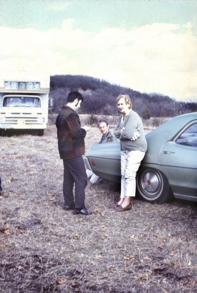 Irene "Granny" York, the owner of York Farm, the location of the Sound Storm music festival, leans on an automobile near two unidentified men, possibly festival staff. There is a camper trailer in the background.