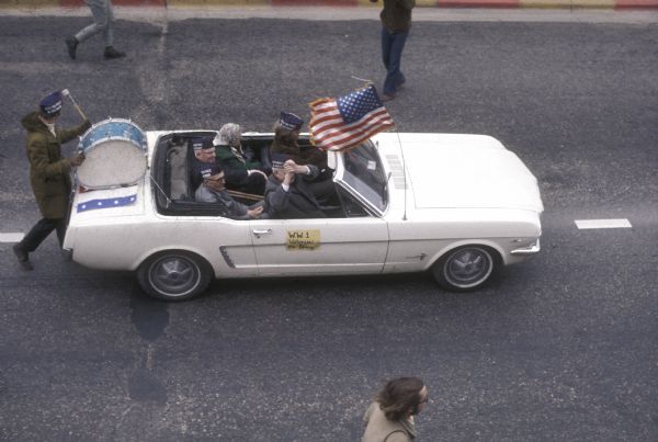 Elevated view of a Ford Mustang carrying four elderly men and one woman driving down a street in a demonstration against the war in Vietnam. An American flag is mounted on the antenna. A sign on the passenger side of the car reads "W.W. 1 Veterans for Peace." Four of the passengers are wearing garrison style hats with text reading "Vets For Peace In Vietnam," and a bass drum is mounted on the trunk with a drummer walking behind the car.