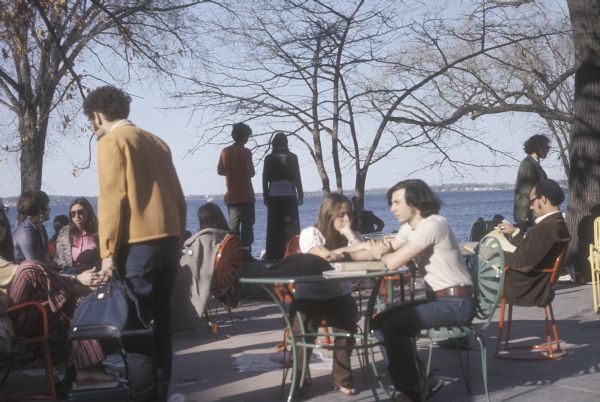 A group of people sitting at brightly colored tables and chairs on the University of Wisconsin Memorial Union Terrace. Lake Mendota is visible in the background.