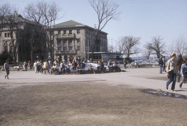 Students sitting on the covered Hagenah Fountain on Library Mall on the University of Wisconsin-Madison campus. The Memorial Student Union and Lake Mendota are visible in the background.