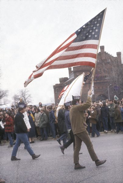Anti-war demonstrators wearing garrison style hats reading "Vietnam Vets For Peace" march in parade carrying American flags and placards while a large crowd watches. The University of Wisconsin-Madison Armory (Red Gym or Old Red) is visible in the background.