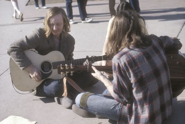 Geoff Cook, wearing suit jacket, and other unidentified individuals, sit on the ground at library mall playing guitars.