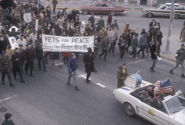 Elevated view of large crowd with a banner reading: "VETS for PEACE / Bring the Troops Home Now." The crowd is following a Ford Mustang carrying five elderly individuals in a demonstration against the war in Vietnam. There is a flag mounted on the antenna of the car, and a sign on the passenger side door reads "W.W. 1 Veterans for Peace." Four of the passengers and many of the crowd members are wearing garrison style hats with text reading "Vets For Peace In Vietnam." A bass drum is mounted on the trunk with a drummer walking behind the car.