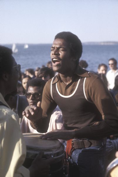 An African-American man plays a djembe-like drum, with other people relaxing in the background on the University of Wisconsin-Madison Memorial Union Terrace. Lake Mendota is visible in the background.