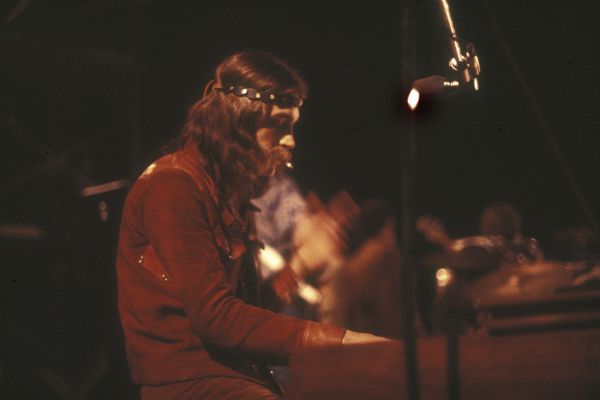 An unknown keyboardist, wearing a headband over long hair and smoking a cigarette, is playing keyboards while performing on stage at night at the Sound Storm music festival.