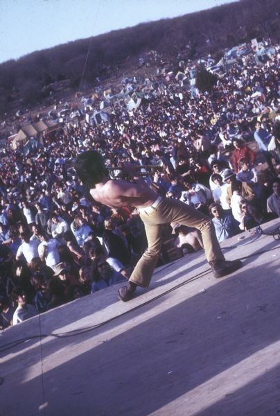 An unidentified trumpeter, shirtless and wearing bell-bottoms, performing at the front of the stage at the Sound Storm music festival, as seen from the rear of the stage looking towards the audience.