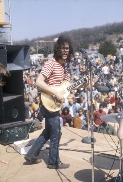 Bob Schmidtke, guitarist for the band Captain Billy's Whiz Band performing with a Gibson Les Paul "goldtop" style guitar on stage at the Sound Storm music festival. The audience in front of the stage, and the camping area on a hill are visible in the background. Schmidtke later joined the group The Tayles who also appeared at Sound Storm.