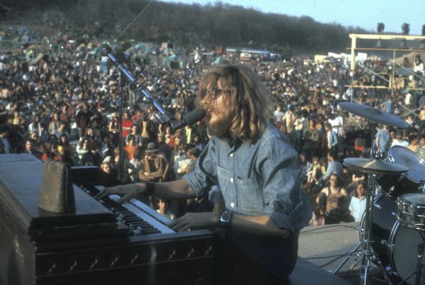 View from stage of Boyd Sibley, keyboardist for the band HOPE, performing onstage at the Sound Storm music festival. There is a cowbell on top of the Hammond "D" and a large crowd is in front of the stage. The campground area on the hill can be seen in the background.