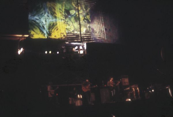 View from audience of unidentified band performing on stage at night at the Sound Storm music festival. There is a large screen above the band with a psychedelic light show projected onto it.