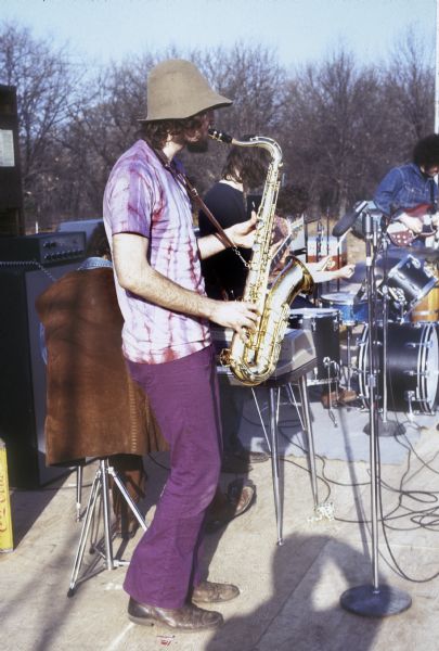 View from side of stage of Rich Keane, wearing a felt bucket hat and a tie-dyed shirt, performing with Strophe band at the Sound Storm music festival. Stewart Weltman is in denim jacket in the background.