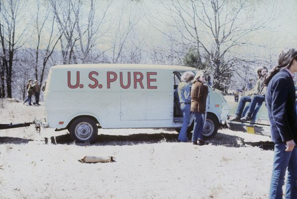 The touring van with trailer for US Pure parked near the backstage area of the Sound Storm music festival. Two people standing outside the van ere roadies and have been identified as Terry Rother and Rich George. They are standing near the open passenger side door, and other people are scattered around among the trees in the background.