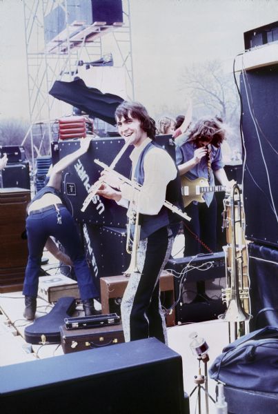 Don Richardson in front the US Pure sound system, grins while holding two flutes and a trumpet, while stagehands unload equipment backstage at the Sound Storm music festival. Richard Wiegel, who is holding a guitar, is standing behind Richardson.