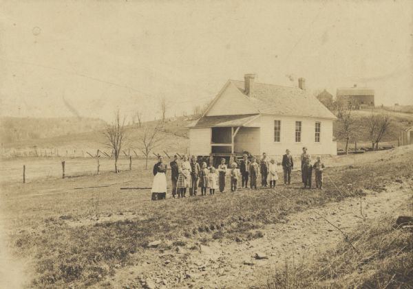 Small white clapboard schoolhouse in South Lancaster Township, with class standing outside in front. There is a barn on a hill in the background.
