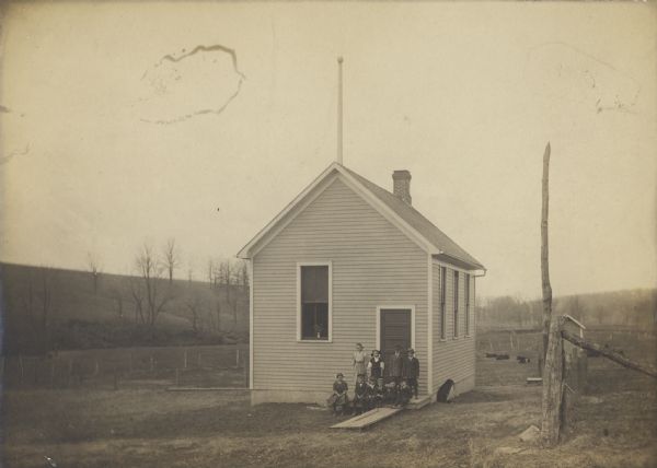 Small rural clapboard schoolhouse with class posing at entrance. There is a dog sitting on the right side of the building, and cows are in a field in the background. On the roof is a chimney, and what appears to be either a flagpole or lightning rod.