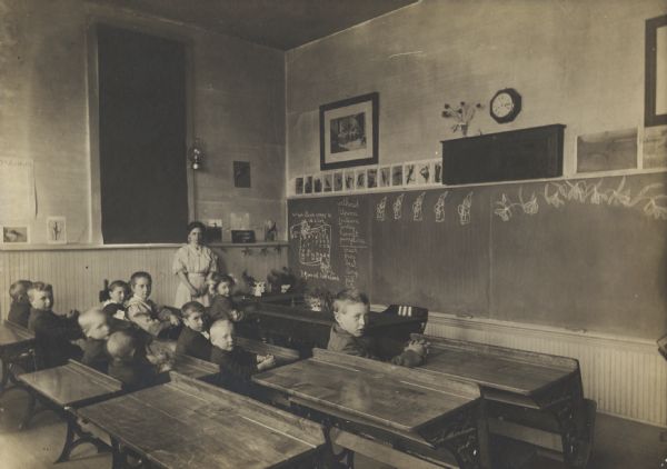 Interior of classroom with teacher and young students, seated in desks. Teacher is identified as Sylvia Rottiger.