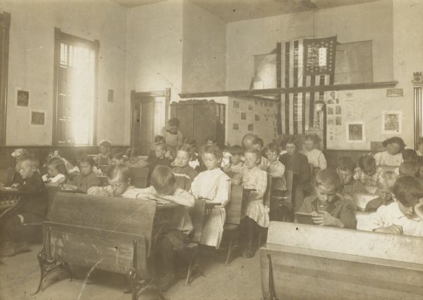 Interior of rural schoolroom. A teacher at the back of the room appears to be helping as students of all ages sit working at their desks. A large U.S. Flag hands in a window.