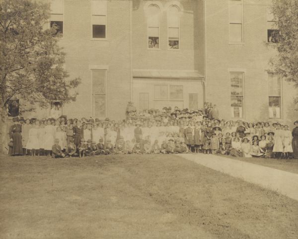 Large group of children and adults posed on the lawn outside a large brick building. Some students are posing in open windows.