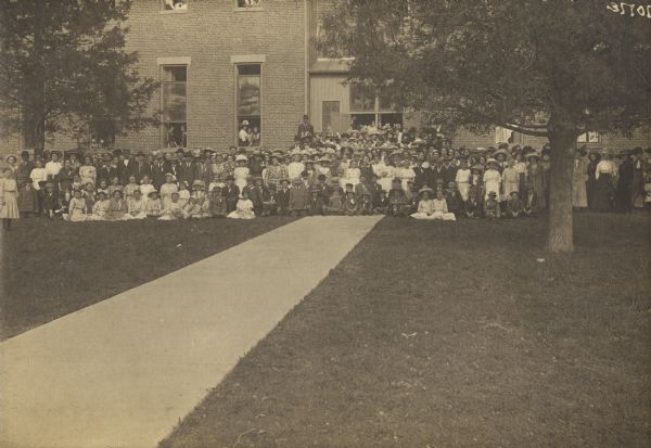 Large group of children and adults posing outside a brick building. Some students are posing in open windows.