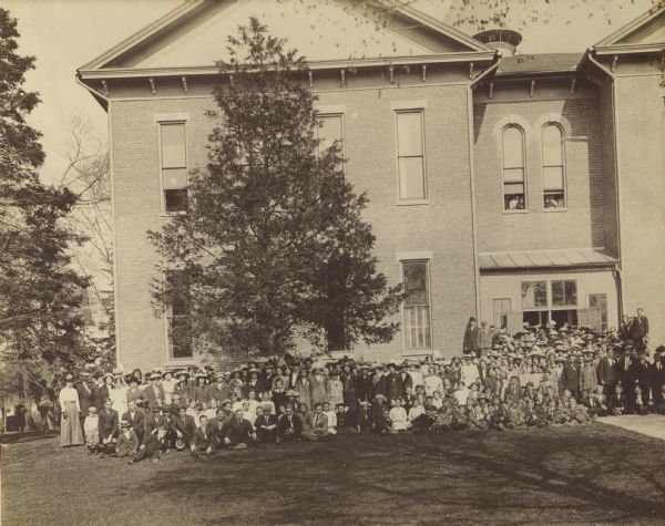Large group of unidentified children and adults pose outside a large brick building. Some students are posing in open windows.
