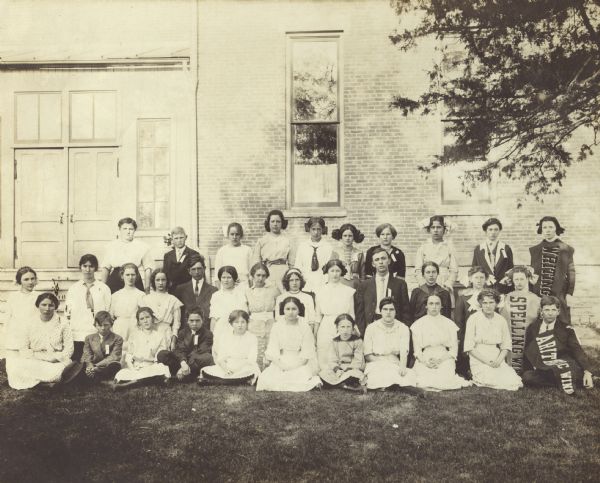Unidentified children wearing ribbons on the lapels or collars pose on the lawn outside a brick building. Some of the children wear banners that say "Spelling Win...," "Writing," and "Athletic Winn..."