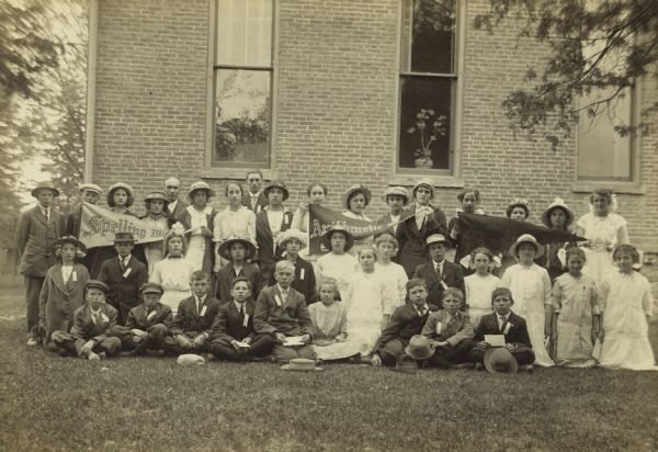 Unidentified group of spelling and arithmetic contest winners posing on a lawn outside of a brick building. The children are wearing ribbons on their lapels or collars, and some of them are holding up banners which read: "Spelling Wi..." and "Arithmetic..."