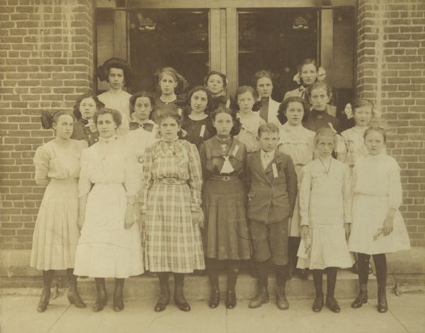 Contest winners pose in doorway of brick building. The children are wearing ribbons on their lapels or collars.
Arithmatic winners, first row from left to right: Bessie Divall, Viola Spiegelberg, Elsie Barr, Florence Wieland, Willie Wieland, Frieda Wetter, Helen Hunter.
Spelling winners, second row: Clara Renalde, Hanna Bast, Florence Uppena, Eva Russell, Susie Walz, Florence Peat, Gladys Elton.
Writing winners, third row: Vina Sedbrook, Orah Brun, Irene Millin, Mary Schauf, Anna Jones