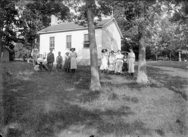 Teacher and students pose outside clapboard one-room schoolhouse. There appears to be a bell tower on the roof.