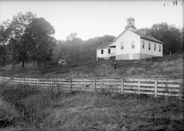 A man stands behind a fence and in front of a clapboard schoolhouse and outhouse on a hill. There is a bell tower on the school building.