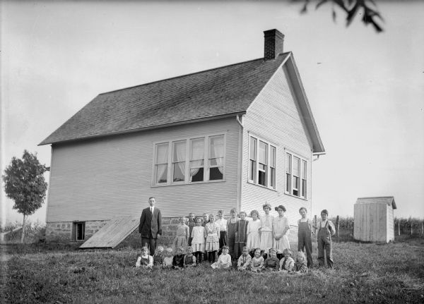 Group portrait of children and their teacher posing on the lawn outside a clapboard school building. On the left side of the building is a stone foundation with a bulkhead.