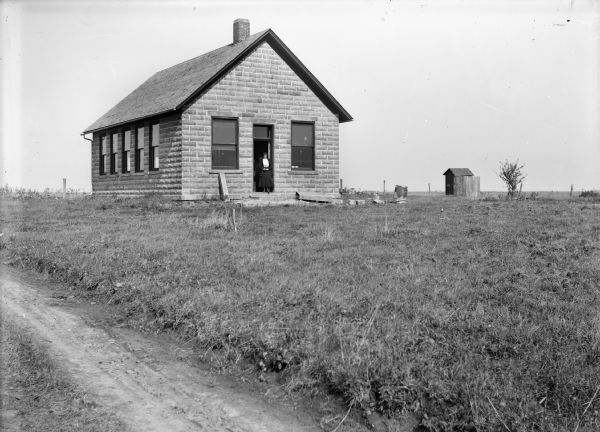 View from dirt road of a woman standing in the doorway of a stone one-room schoolhouse. An outhouse is behind the school.