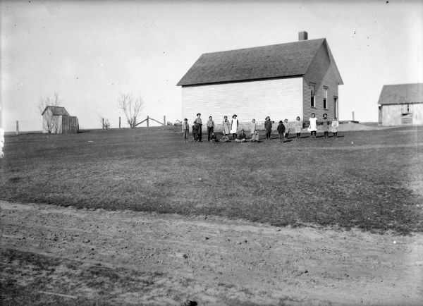 Teacher and students posed on lawn in front of clapboard school building. An outhouse and shed are behind the building on the right and left.