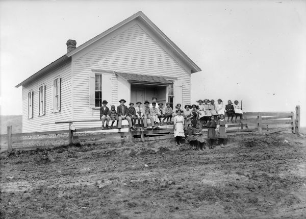 Group of students sitting and standing along fence in front of a clapboard one-room school. The teacher stands behind them near the front door which has roof over the entrance and side walls.