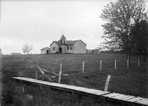 View over wooden bridge of school building. There is a ladder near the front entrance which has a small bell tower. There is  a shed on the left, and an outhouse and a fence on the right.