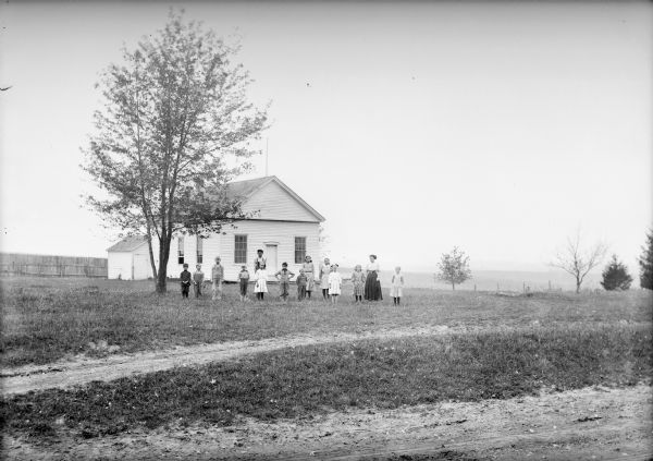 Teacher and students posing on grounds in front of a clapboard one-room schoolhouse. There is a fence and outhouse on the left, and in the background is a field.