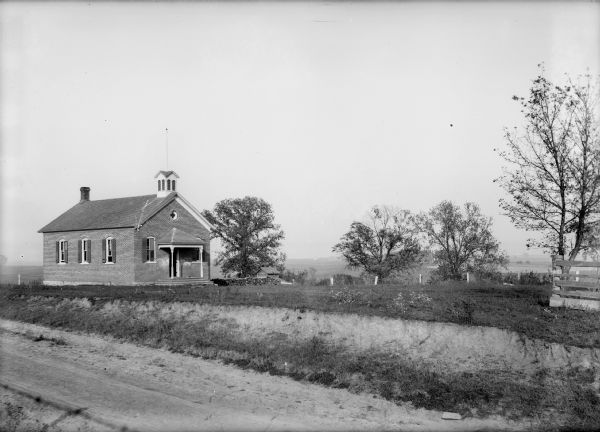 View from dirt road of brick schoolhouse with porch and small bell tower.
