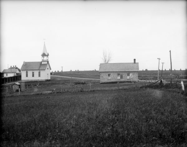 View across field of stone school building with outhouse and a wood pile. In the background is a church with a belfry. A road is along the right leading up to a hill and other buildings.