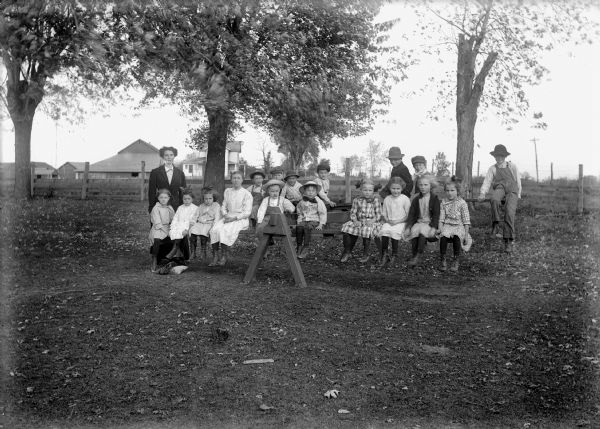 Students and teacher pose outdoors. The students are sitting on two teeter totters. In the background is a fence and beyond is a farmhouse and farm.