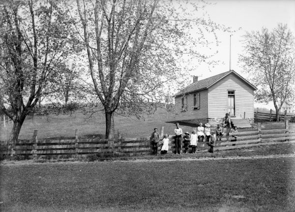 Jamestown students sitting and standing along a fence in front of clapboard one-room school building on a small hill.