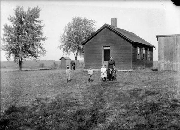 View of a teacher and four students standing in front of a clapboard one-room school building. A horse is grazing in the grass in front of an outhouse on the left. There is a shed on the right.