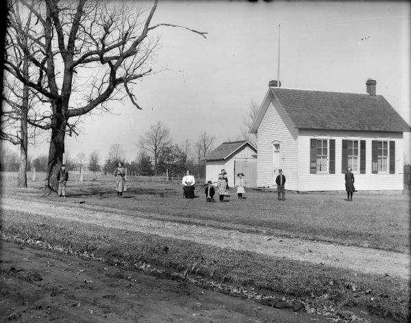 View from dirt road or field of a teacher sitting and students posing in the yard in front of a clapboard one-room school building.