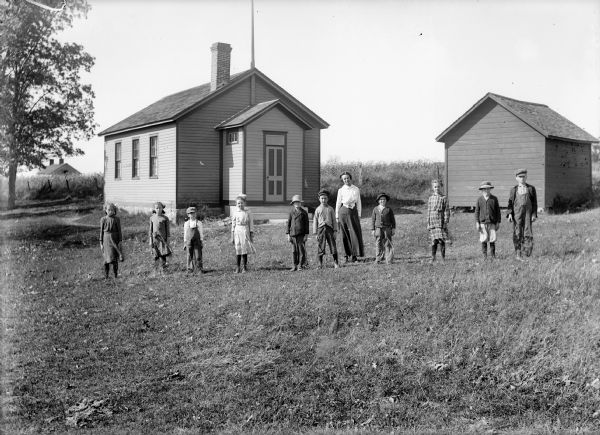 Teacher and students stand spaced apart in a line in front of a clapboard one-room school building and a small shed, perhaps an outhouse. In the background on the left is a field and another building.